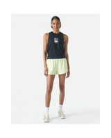 A model stands against a plain white studio background wearing a sleeveless black athletic tank top with a white logo on the chest, light yellow running shorts, white socks, and metallic sneakers.