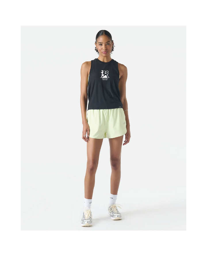 A model stands against a plain white studio background wearing a sleeveless black athletic tank top with a white logo on the chest, light yellow running shorts, white socks, and metallic sneakers.