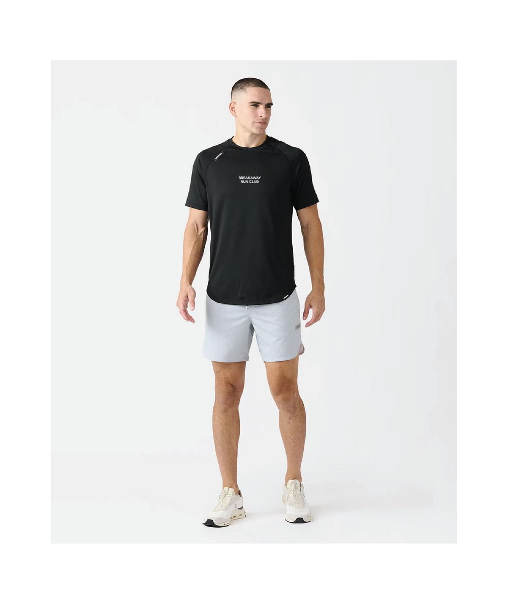 A man modeling a black short-sleeve athletic t-shirt with white text on the chest, paired with light gray running shorts and white sneakers, against a plain white studio background.