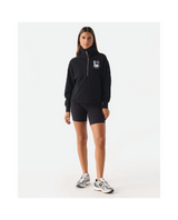 A woman modeling a black half-zip pullover hoodie with a white chest logo, paired with black athletic shorts and white sneakers, against a plain white studio background.