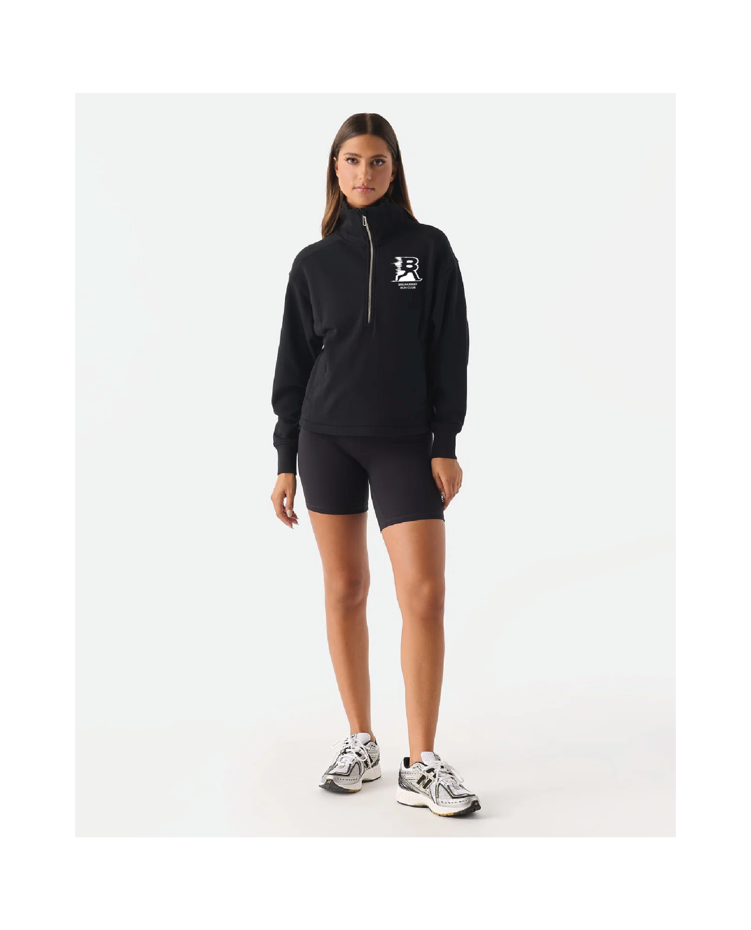 A woman modeling a black half-zip pullover hoodie with a white chest logo, paired with black athletic shorts and white sneakers, against a plain white studio background.
