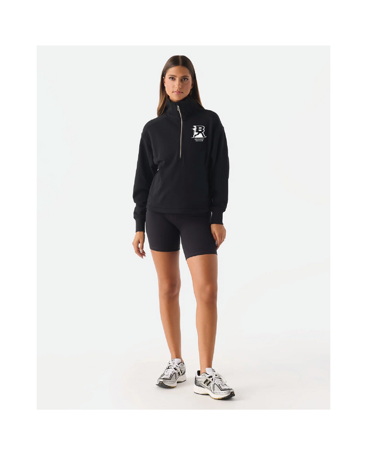 A woman modeling a black half-zip pullover hoodie with a white chest logo, paired with black athletic shorts and white sneakers, against a plain white studio background.