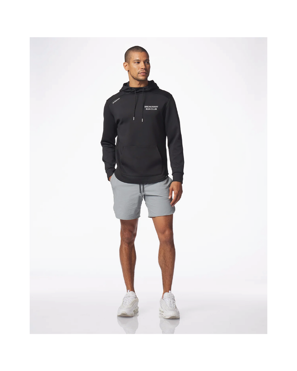 A man modeling a black pullover hoodie with white drawstrings and a small white chest logo, paired with light gray athletic shorts and white sneakers, against a clean white studio background.