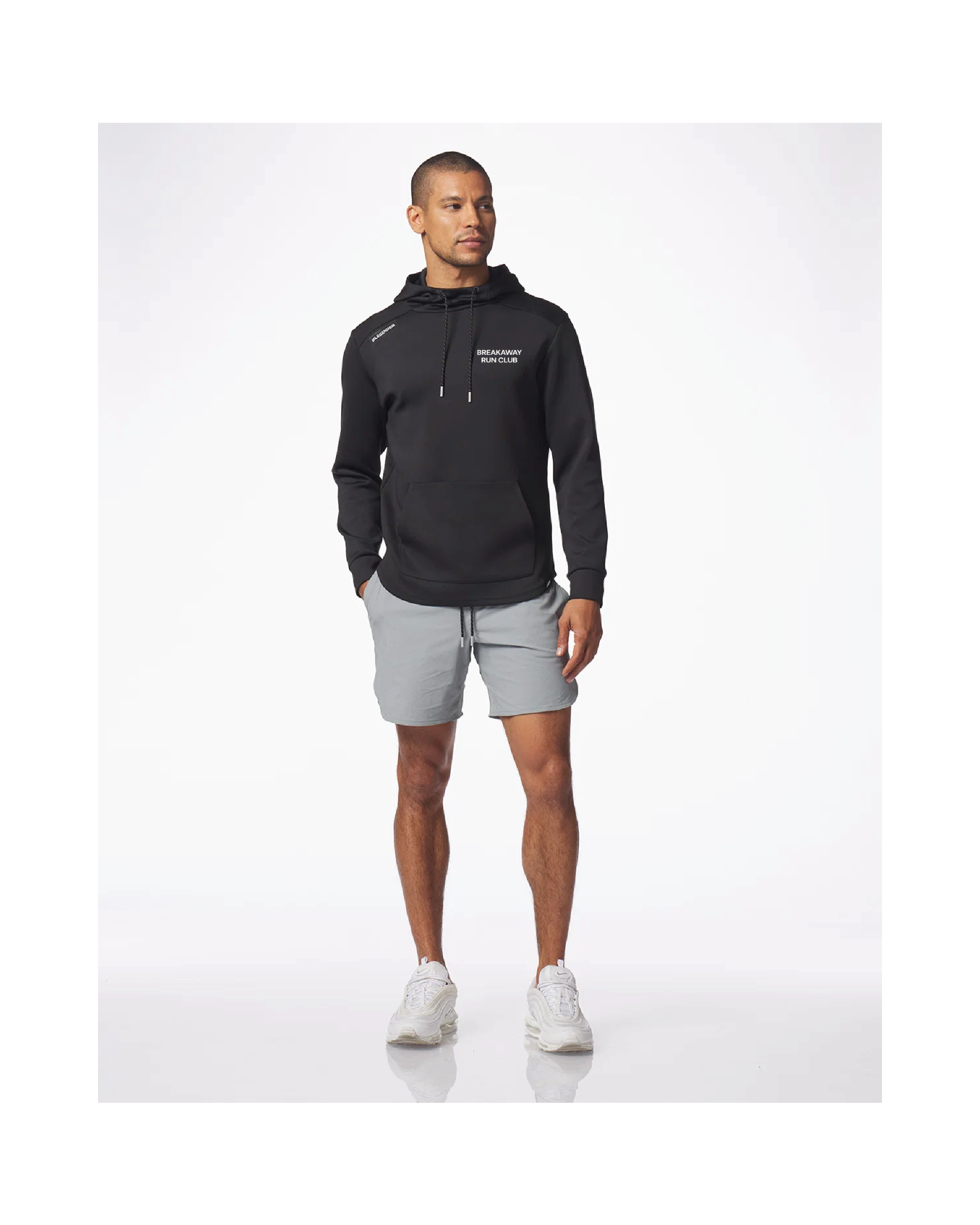 A man modeling a black pullover hoodie with white drawstrings and a small white chest logo, paired with light gray athletic shorts and white sneakers, against a clean white studio background.